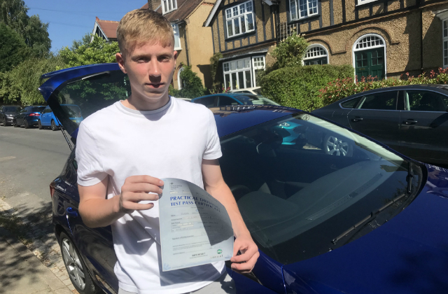 Finbar King holding his driving test pass certificate while standing in front of a blue car with its boot open.