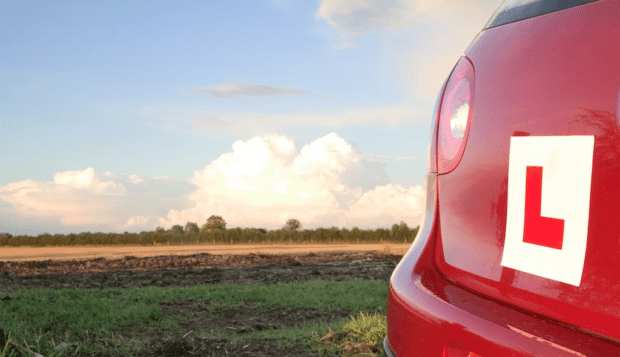 The rear of a red car with L plates, with a view of the countryside in the background. 